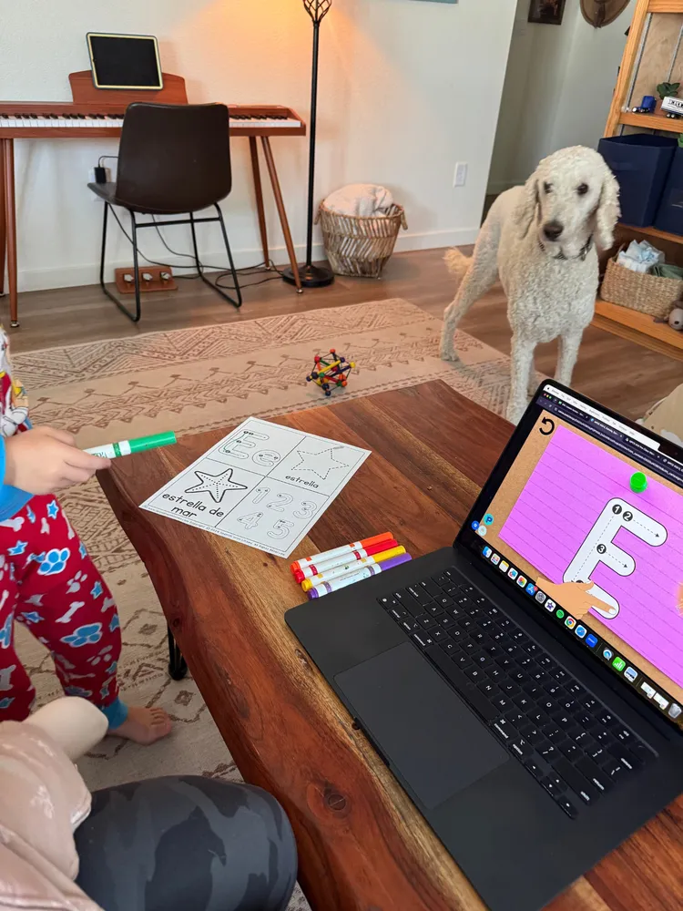 A little one follows a Spanish lesson on a laptop at the coffee table, with a worksheet, markers and the family dog nearby