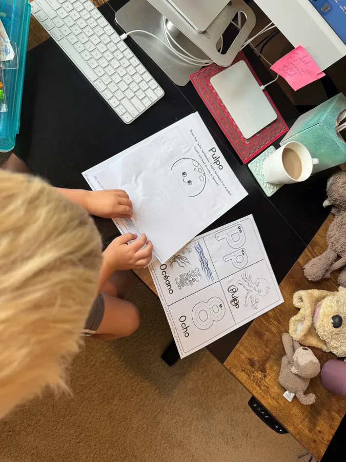 Seen from above, a child traces a Pulpo worksheet at a desk beside a keyboard and stuffed animals